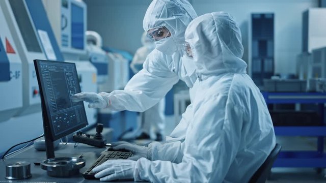 Factory Cleanroom: Engineer and Scientist Wearing Coveralls and Masks Have Discussion, Use Computer Showing Infrastructure System Control. In Background CNC Machinery, Robot Arm, Electronics Equipment