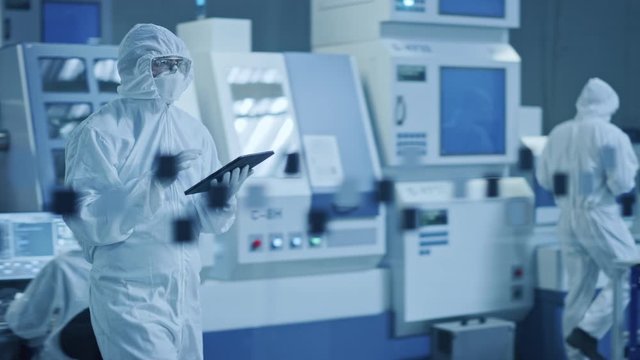 Research Factory Cleanroom: Engineer Wearing Coverall and Mask, Walking Through Workshopop Uses Tablet Computer. In Background Professionals Work on Modern High Tech CNC Machinery