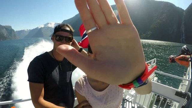 Couple on a fjord boat tour with a Norwegian flag waving behind them, Songefjorden Norway. The motor of the ship makes the water wavy and foamy. Lush green mountains surrounding the fjord. Strong wind