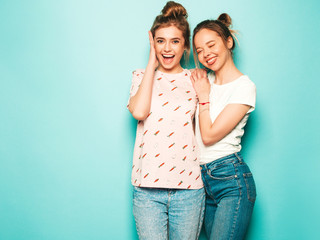 Two young beautiful blond smiling hipster girls in trendy summer hipster jeans clothes. Sexy carefree women posing near blue wall. Trendy and positive models having fun