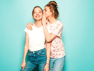 Two young beautiful blond smiling hipster girls in trendy summer hipster jeans clothes. Sexy carefree women posing near blue wall. Trendy and positive models having fun. Female shares secret, gossip