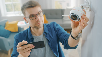 Young Man in Glasses Wearing a Blue Shirt is Adjusting a Modern Wi-Fi Surveillance Camera with Two...
