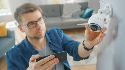 Young Man in Glasses Wearing a Blue Shirt is Adjusting a Modern Wi-Fi Surveillance Camera with Two Antennas on a White Wall at Home. He's Checking the Video Feed on his Smartphone.