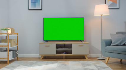 Shot of a TV with Horizontal Green Screen Mock Up. Cozy Living Room at Day Time with a Chair and Lamps Turned On at Home.