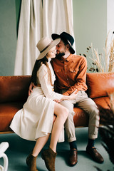 Obraz premium stylish couple in love with hats sitting on a brown sofa, hugging in a spacious beautiful Studio
