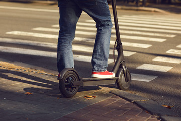 Legs of a man in jeans and sneakers on an electric scooter at a crosswalk on a city street