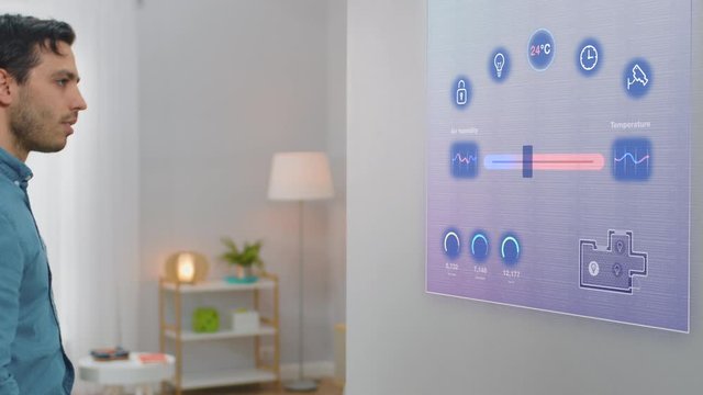 Handsome Young Man Interacts with Augmented Reality Display. He is Changing Climate Control Setting to a Higher Temperature. Smart Home Concept with Transparent Screen Made with VFX Special Effects.