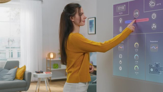 Beautiful Young Female Interacts with Augmented Reality Display. She's Changing Climate Control Setting to Higher Temperature. Smart Home Concept with Transparent Screen Made with VFX Special Effects.