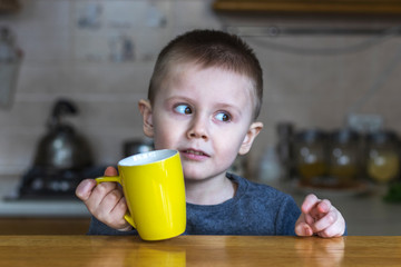 Portrait of pretty little boy in the kitchen at home. Surprised child looks away. Child sits at the kitchen table and holds a yellow cup in his hand.