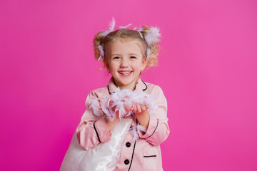 Studio shot of a contented, beautiful little girl posing in pink pajamas. Funny baby girl in pyjamas standing on a pink background with a pillow, feathers and a clock