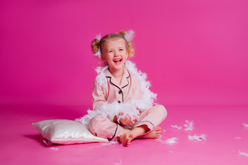 Studio shot of a contented, beautiful little girl posing in pink pajamas. Funny baby girl in pyjamas standing on a pink background with a pillow, feathers and a clock