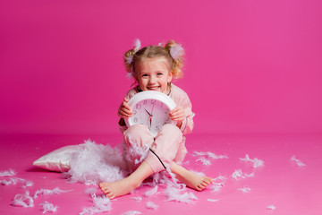 Obraz na płótnie Canvas Studio shot of a contented, beautiful little girl posing in pink pajamas. Funny baby girl in pyjamas standing on a pink background with a pillow, feathers and a clock