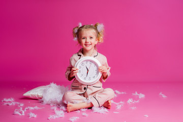 Studio shot of a contented, beautiful little girl posing in pink pajamas. Funny baby girl in pyjamas standing on a pink background with a pillow, feathers and a clock