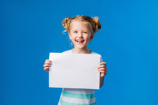 baby girl holding a white sheet.Cute little girl with a white sheet of paper.blue background.space for text.A little girl holds an empty piece of paper