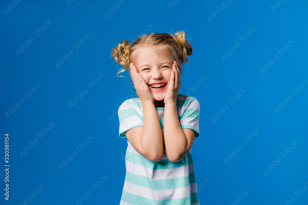 Wall mural Cute little girl blonde smiling holding hands on sides standing on a blue background, happy child on a blue background, space for text