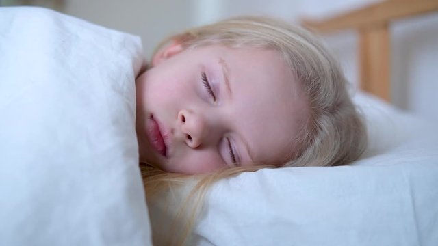 Sleeping little blonde girl close up in big bed with white bedding early in the morning. Children dream. Childhood, calm, night .4k footage
