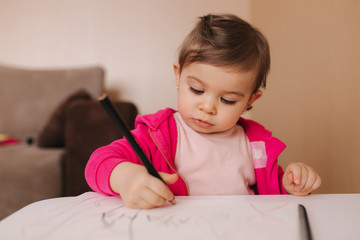 Naklejka premium Happy baby girl sit at the table and wthire something. Little girl use pencil for drawing on white paper at home