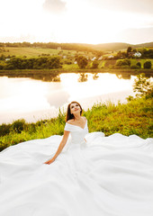 Fototapeta na wymiar Charming bride in a lush white dress sits on the grass against a lake background