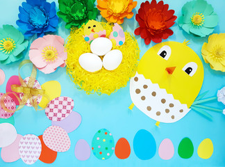 Child made crafts his own hands at Easter. Handmade nest with eggs, flowers, chicken, chicken in an egg, frame of eggs. Cute art creativity on blue background. Flat lay, top view, close up, copy space