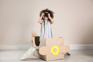 Cute African American child playing with cardboard plane and binoculars near beige wall