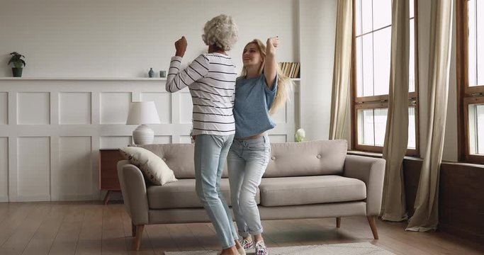 Full length excited elderly mature woman listening to favorite music, dancing with grown up daughter at home. Happy bonding two generations family having fun laughing enjoying energetic activity.