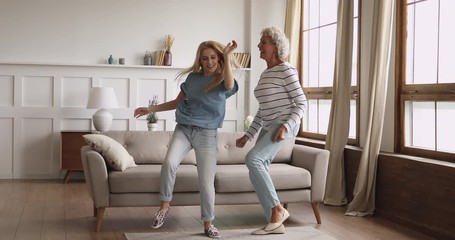 Full length overjoyed young lady dancing with excited middle aged elderly mother. Happy two generations family having fun together in modern living room, enjoying energetic activity, jumping at home. - Powered by Adobe