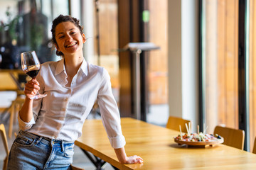 Portrait of one young beautiful caucasian woman standing by the table leaning holding glass of red wine at home alone smiling wearing white shirt looking to the camera front view