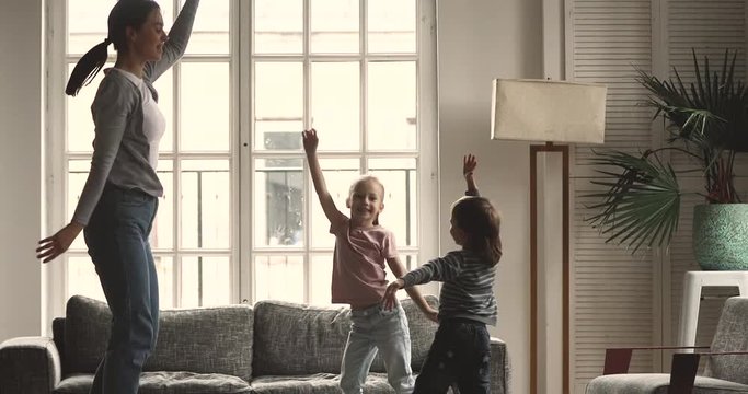Active young mommy nanny dancing to favorite music, jumping with little cute son and daughter in living room. Energetic happy small kids siblings having fun with millennial mother babysitter at home.