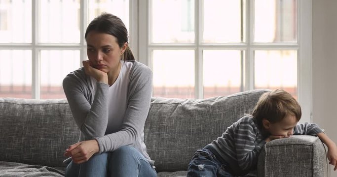 Stressed young woman sitting separately on sofa with sad little preschool boy, annoyed by bad kids behavior. Nervous worried millennial mother babysitter ignoring bored small child son at home.