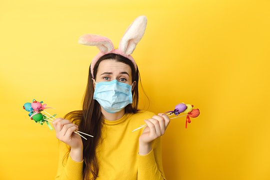Puzzled young woman with virus mask, sad to celebrate Easter alone, holds many colored eggs, wears fluffy earsr , isolated on yellow background. Concept holiday, N1H1 coronavirus, virus protection