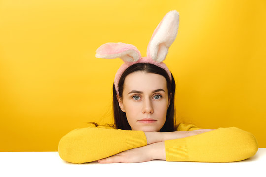 Image of lovely female leans on white table, wears fluffy ears, dressed in sweater, models over yellow background with copy space. People, special holiday, celebration, Easter concept