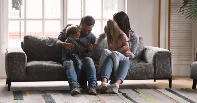 Full length happy young married couple relaxing on sofa, tickling little cute children siblings. Overjoyed family of four playing, parents having fun together with small kids in modern living room.