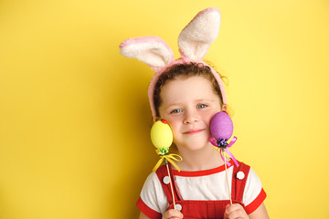 Obraz na płótnie Canvas Portait of cheerful curly little girl wears bunny ears, holds colored Easter eggs, likes celebration and mystery of holiday, dressed in t-shirt, isolated on yellow background. Seasonal holiday concept