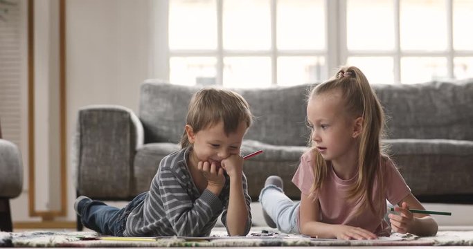 Small cute child boy lying on floor carpet with adorable elder sister, drawing together in paper album. Little kids siblings having fun, enjoying hobby pastime alone without parents in living room.