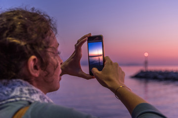 Blonde woman taking a photo with her mobile phone in front of a lake during sunset