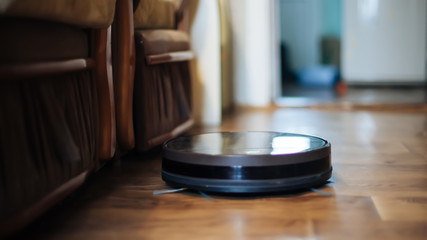 robot vacuum cleaner removes dust in room on brown floor. vacuum cleaner in ordinary apartment near chairs. modern household wireless device for cleaning house