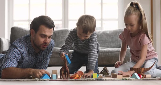 Loving single father lying on floor with adorable little son and daughter, playing with toys together in living room. Happy young daddy enjoying free weekend leisure playtime with children siblings.