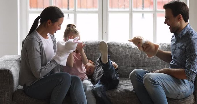 Happy young couple parents wearing puppet hand toys, tickling small laughing son and daughter. Overjoyed full family of four having fun on comfortable couch in living room, enjoying weekend playtime.