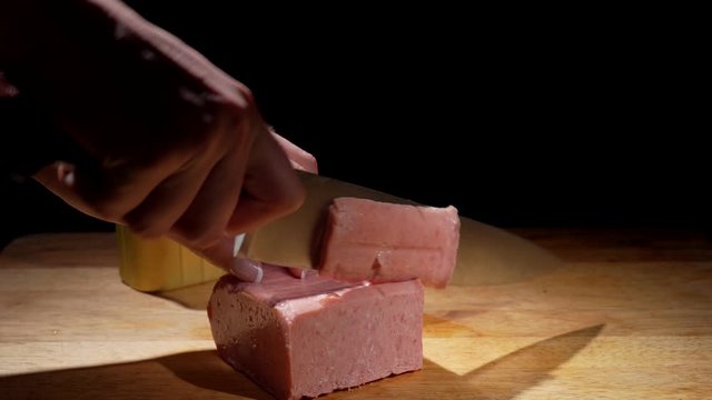 This video shows anonymous hands chopping and cutting canned ham on a wooden cutting board with a black background.