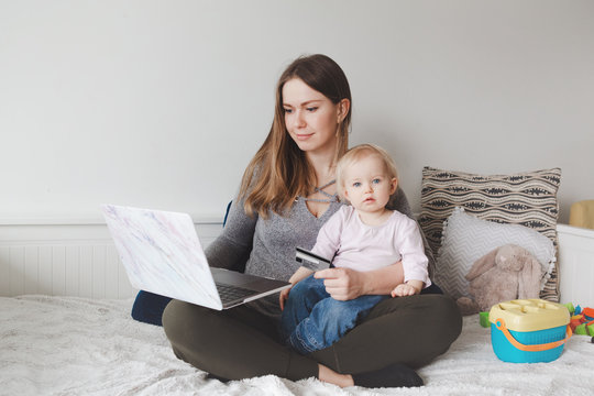 Young Caucasian Mother With Baby Shopping Online From Home. Stay At Home Single Mom With Kid Toddler Daughter Buying Ordering Products Food On Internet And Paying With A Credit Card.