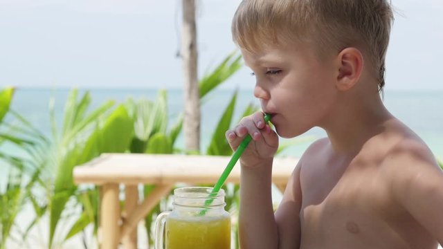 little boy on the beach drinks juice through a straw. Portrait of a little boy. A child sits at a table in the park and drinks juice through a straw. Bright sunny day in the summer.