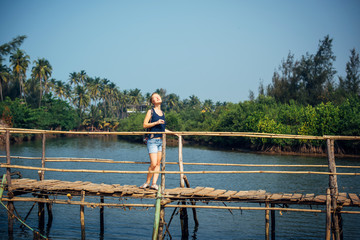 A female tourist in denim shorts with short haircut on sunny tropical day. Young pretty girl stands on wooden bridge over small river against palm jungle and blue sky. Trips to picturesque places.