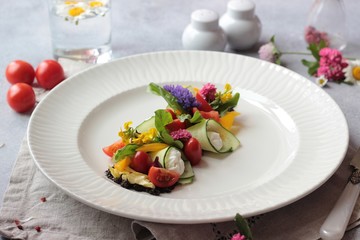 Colorful vegetable salad decorated with flowers on a white plate, light grey stone background. Healthy spring or summer lunch.
