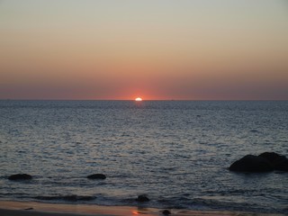 Part of the fiery red disk of the sun peeking out from the horizon. Orange ray and glow of sunset on the expanse of sea. Stones in water and sandy coast on a foreground, the open ocean on a background