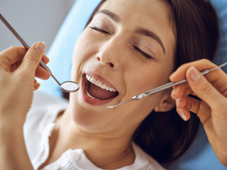 Smiling brunette woman being examined by dentist at dental clinic. Hands of a doctor holding dental instruments near patient's mouth. Healthy teeth and medicine concept