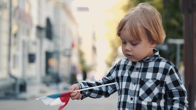 Blond boy holds Czech flag. Kid in checkered shirt is on street. Child is alone in city center.