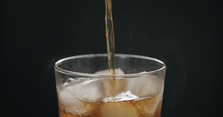 pour cola in glass with ice cubes on black background closeup