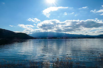 An idyllic view on Mt Fuji from the side of Kawaguchiko Lake, Japan. The volcano is surrounded by clouds. Dried, golden grass on the shore of the lake. Serenity and calmness. Bright and clear day.