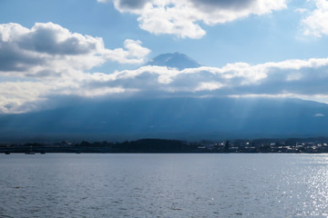 Fototapeta na wymiar A close up view on Mt Fuji from the side of Kawaguchiko Lake, Japan. The mountain is hiding behind the clouds. Top of the volcano covered with a snow layer. Serenity and calmness. Calm lake's surface
