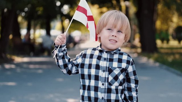 A boy is holding flag of England. He is in park in summer. Kid wears checkered shirt.